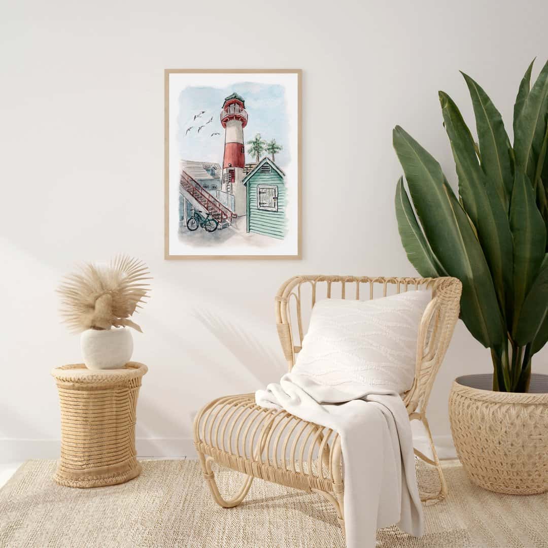Coastal watercolor print of a red and white lighthouse with a green beach house, palm trees, and a bike in front. Perfect nautical wall art for beach-inspired home décor. Framed artwork in a modern boho living room setting with wicker chair and white blanket.