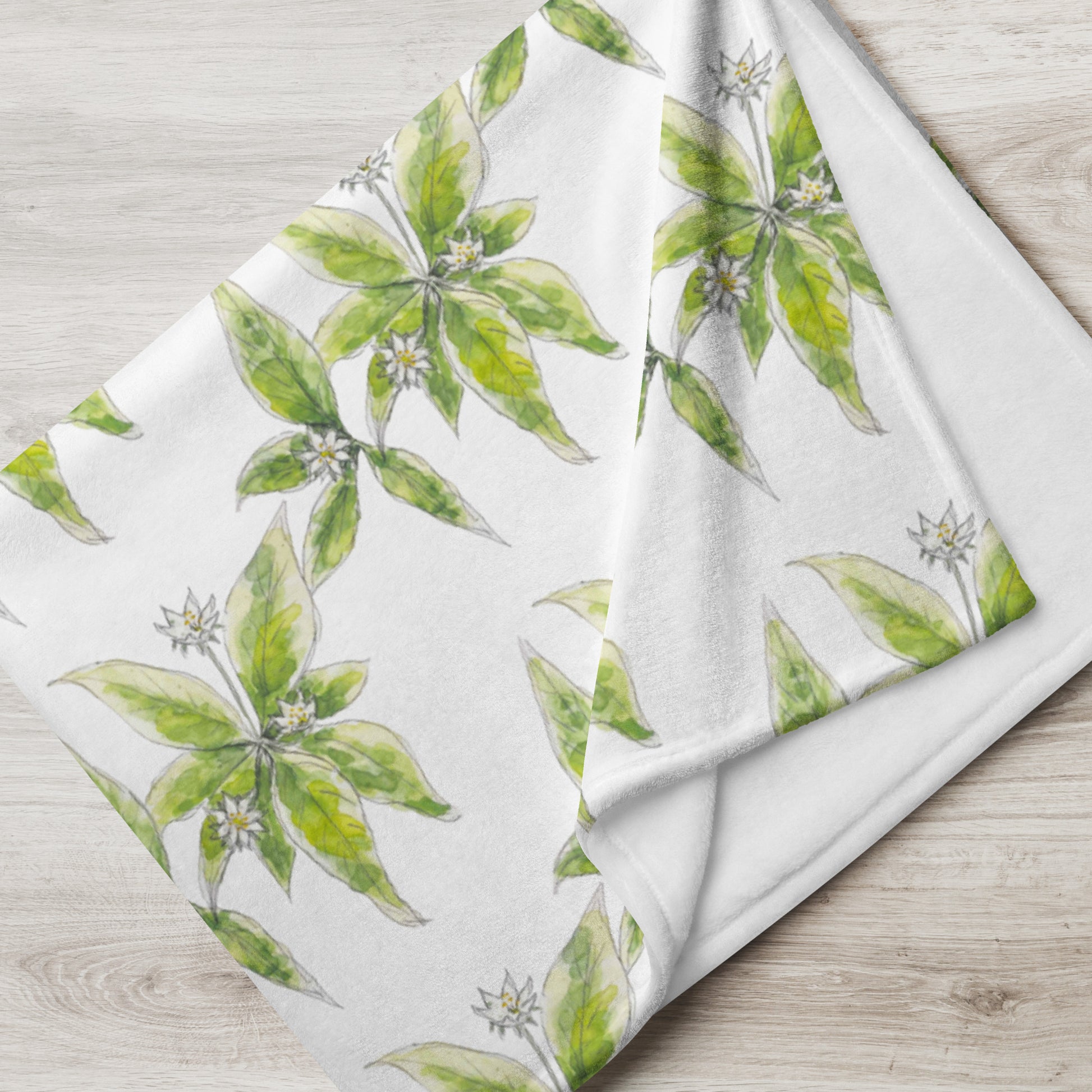 Soft fleece throw blanket featuring a white starflower pattern with green leaves, elegantly folded on a light wooden surface. Cozy botanical home décor with nature-inspired watercolor design.