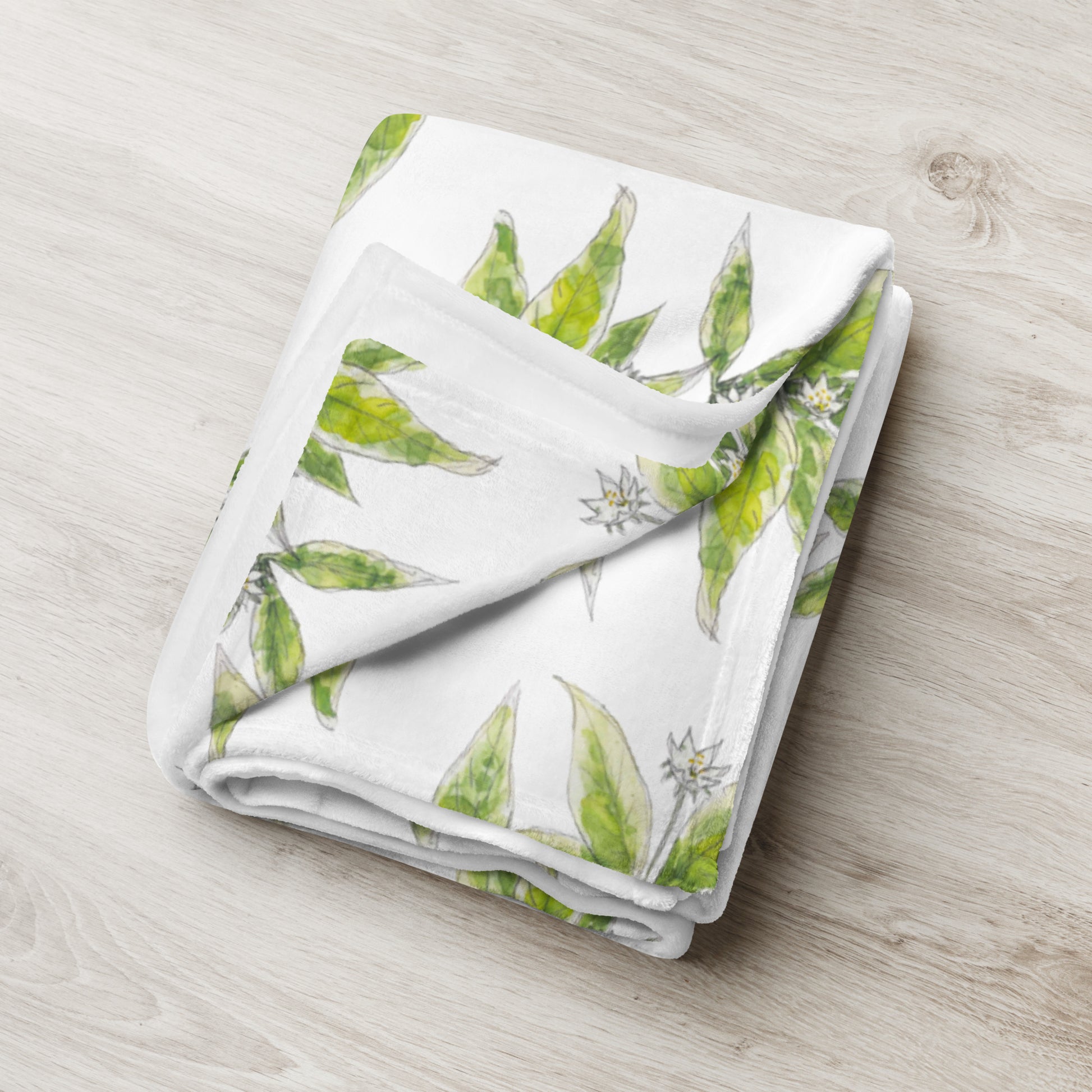 Soft fleece throw blanket featuring a white starflower pattern with green leaves, elegantly folded on a light wooden surface. Cozy botanical home décor with nature-inspired watercolor design.