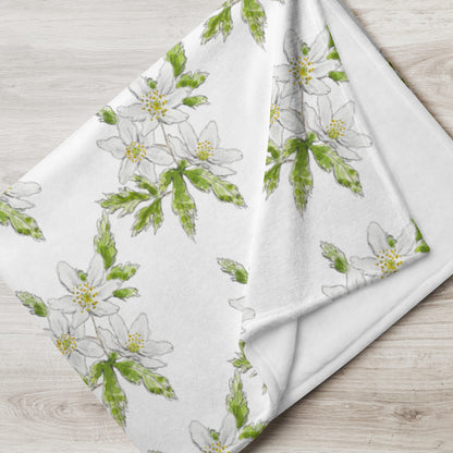Soft fleece throw blanket featuring a white wood anemone flower pattern with green leaves, elegantly folded on a light wooden surface. Cozy botanical home décor with nature-inspired watercolor design.