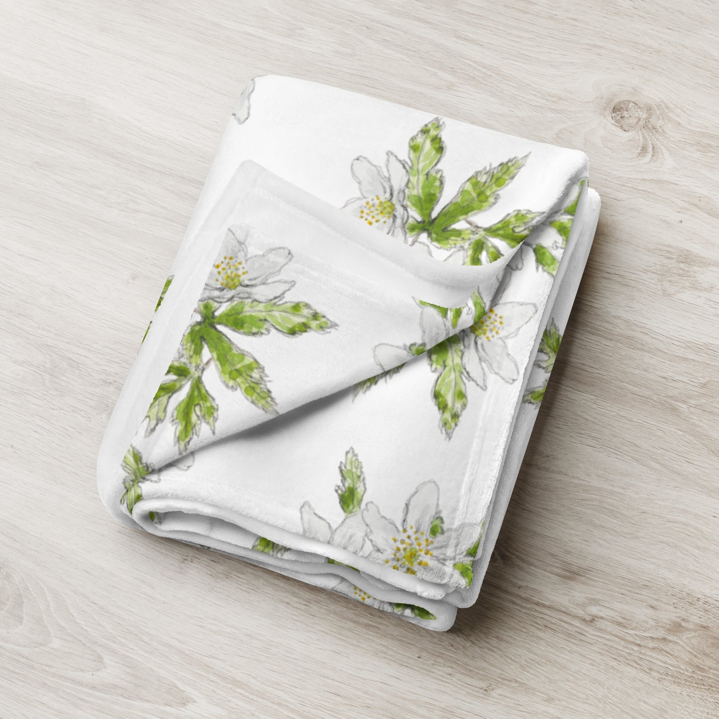 Soft fleece throw blanket featuring a white wood anemone flower pattern with green leaves, elegantly folded on a light wooden surface. Cozy botanical home décor with nature-inspired watercolor design.