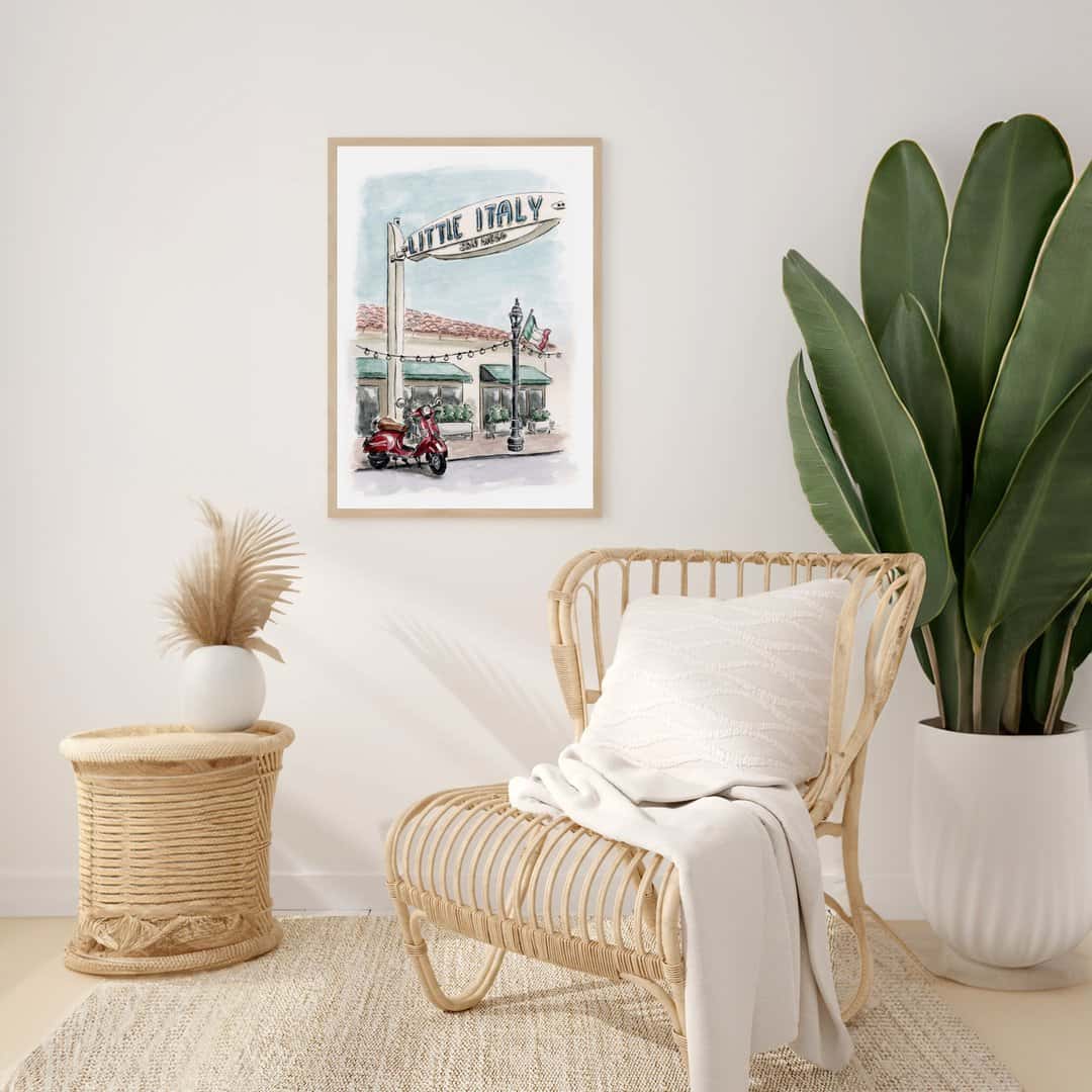 Watercolor print of Little Italy San Diego street scene with red Vespa scooter and iconic Little Italy sign displayed at a modern boho living room setting with wicker chair and white blanket. Perfect artwork for lovers of San Diego and Italian-inspired decor.

