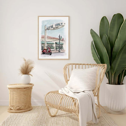 Watercolor print of Little Italy San Diego street scene with red Vespa scooter and iconic Little Italy sign displayed at a modern boho living room setting with wicker chair and white blanket. Perfect artwork for lovers of San Diego and Italian-inspired decor.
