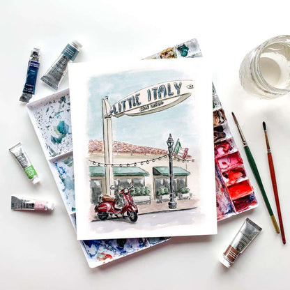 Watercolor painting of Little Italy San Diego with red Vespa scooter, displayed with artist's paint palette and brushes, handmade travel-inspired artwork for modern decor, Italian-themed wall art, perfect for gifts and home decoration
