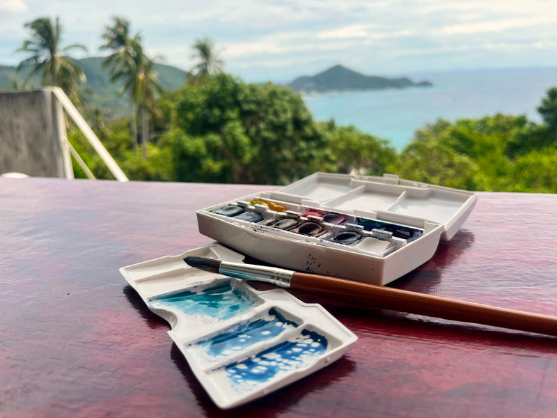 Watercolor paint set with a brush on a table with a scenic view of trees and the ocean in the background