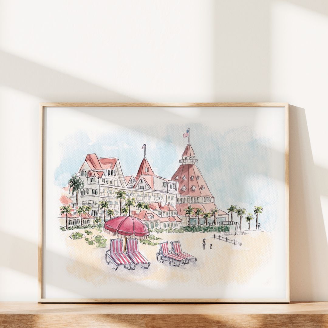 Swil Arts’ The Del’s Timeless Charm is framed California art showcasing the iconic Hotel del Coronado, palm trees, and striped red-and-white umbrellas on the sand, displayed against a sunlit white wall.